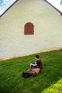 Side view of young man sitting on grass by historic whitewashed stone barn
