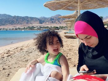 Mother using mobile phone while sitting with daughter at beach