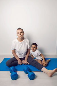 Grandmother and her grandson are sitting on a yoga mat in a white apartment at home