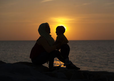 Rear view of couple sitting on beach against sky during sunset