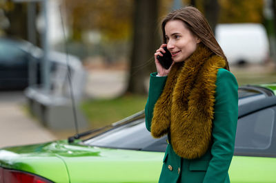 A woman talking on the phone while standing by a green car parked on a city street in the background