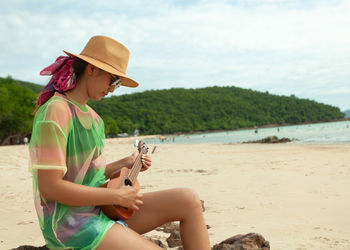 Woman playing ukulele while sitting at beach against sky