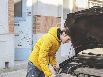 A young caucasian guy in a yellow jacket inspects an internal breakdown with an open car hood