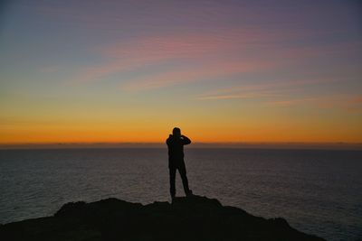 Silhouette man standing on rock at beach against orange sky