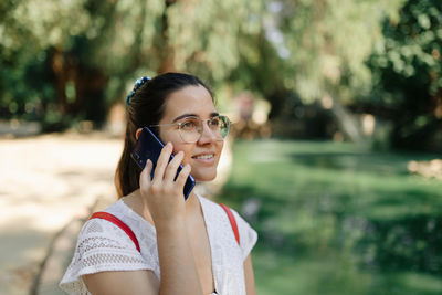 Medium shot of a young woman with a white dress and a red backpack talking on the phone in a park