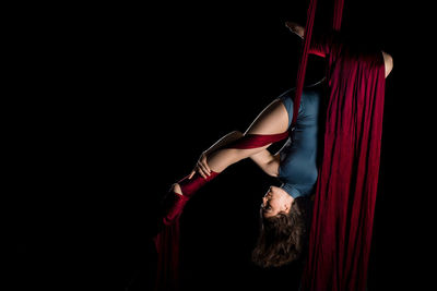 Young woman hanging on fabric while dancing against black background