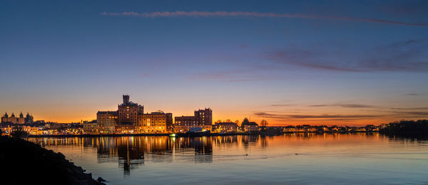 River by illuminated buildings against sky at sunset