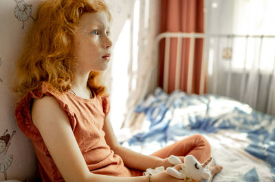 Thoughtful redhead girl with soft toy sitting on bed