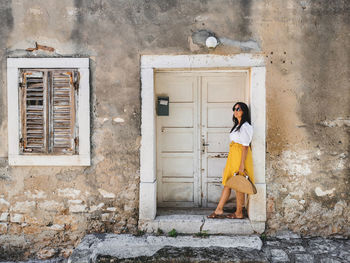 Young woman in summer outfit standing in front of old white wooden door of an old house.