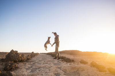 Full length of man playing with dog while standing on land against clear sky during sunset