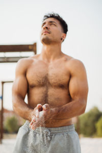 Muscular hispanic ethnic male athlete with naked torso rubbing sand on hands during training on the beach looking away