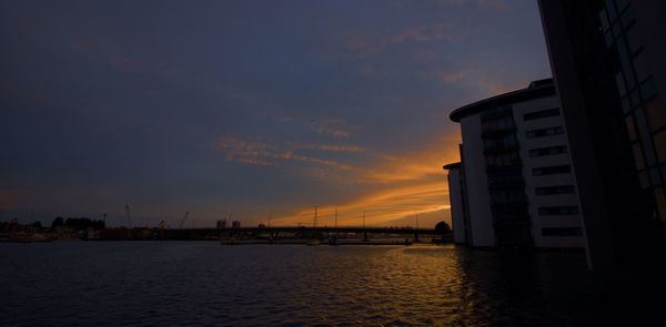 Silhouette buildings by sea against sky at sunset