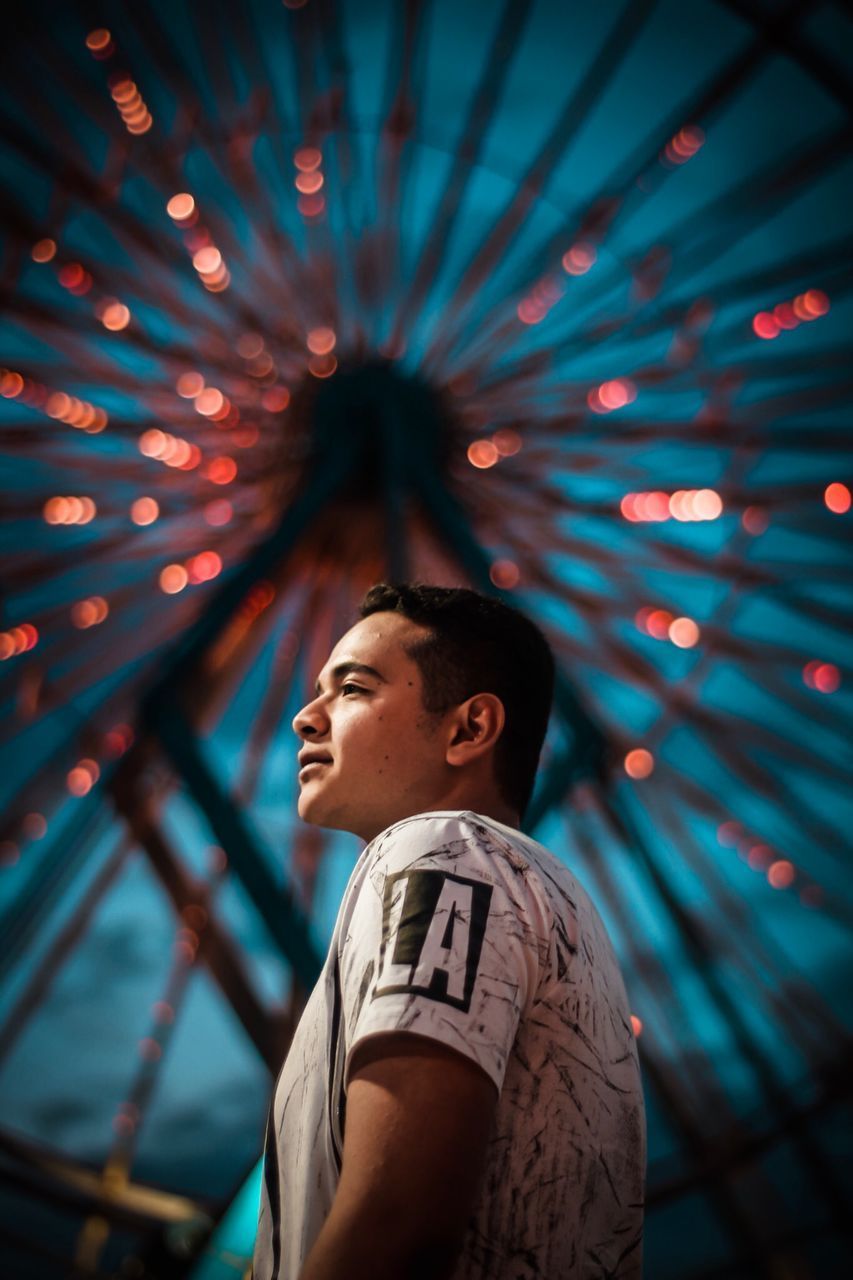 LOW ANGLE VIEW OF YOUNG MAN STANDING AGAINST ILLUMINATED LIGHT