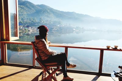 Side view of woman looking at lake while sitting by railing during sunny day