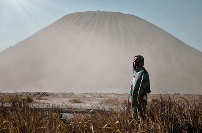 Side view of young man standing on field against mountain during sunny day