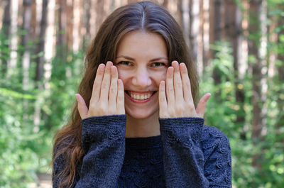 Portrait of smiling young woman holding hands to face while standing against trees