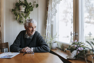 Senior man looking through window while sitting at dining table in home