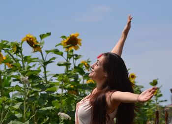 Young woman with arms outstretched standing by sunflowers against sky