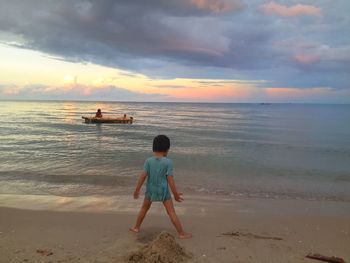 Rear view of boy standing at beach against cloudy sky