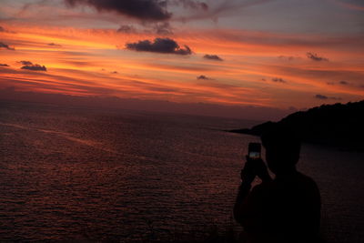 Silhouette person photographing sea against sky during sunset