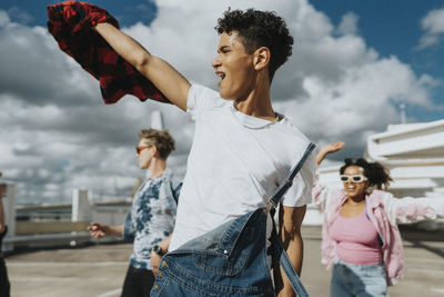 Cheerful hipster man in bib overalls dancing with male and female friends in parking lot