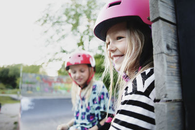 Smiling girl wearing helmet sitting with friend on edge of skateboard ramp