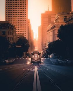 Cars on road by buildings against sky during sunset