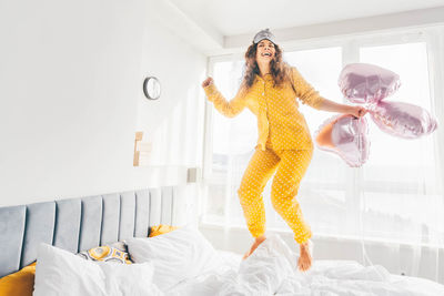 Full length of woman with arms raised standing on bed at home