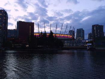 Illuminated buildings by river against sky at sunset