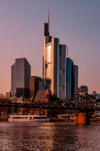 Modern buildings by river against sky during sunset