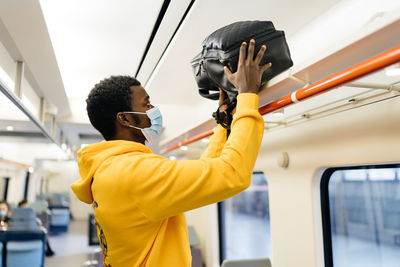 Side view of african american male traveler in medical mask standing in train and putting backpack on shelf before departure