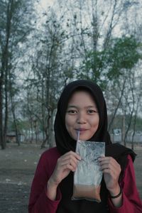 Portrait of young woman having drink while standing against trees