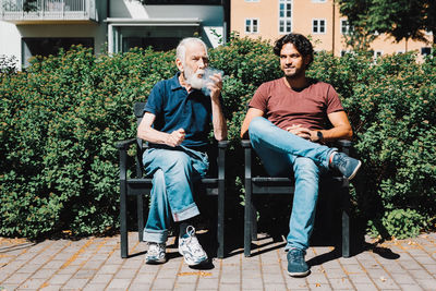 Full length of retired senior man smoking pipe while sitting with male caretaker against nursing home