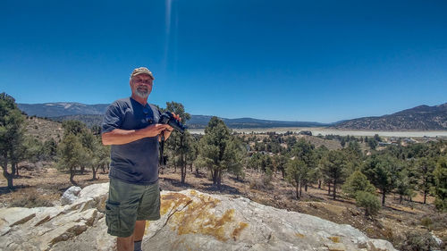 Man holding digital camera while standing on mountain against clear blue sky during sunny day