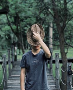 Young man wearing hat while standing on footbridge against trees in forest