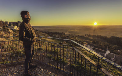 Side view of man looking at landscape while standing by railing during sunset
