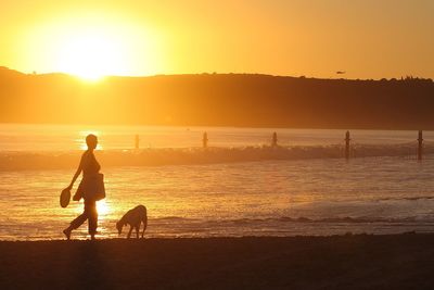 Woman with dog walking at beach against sky during sunset