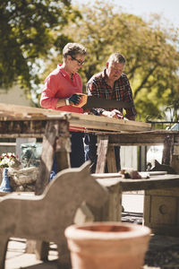 Senior woman cutting wooden plank with saw while working with man in yard