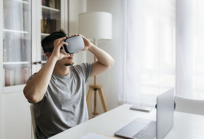 Male entrepreneur holding virtual reality headset while sitting with laptop on desk at home office
