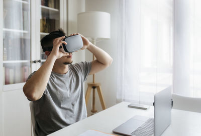 Male entrepreneur holding virtual reality headset while sitting with laptop on desk at home office
