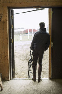 Full length rear view of young man with bridle leaning in doorway of horse stable