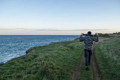 Rear view of man carrying tripod on trail by sea against sky