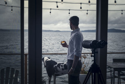 Man enjoying a whiskey on ice cocktail drink while gazing out over an overcast sea with pet dog.