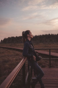 Woman sitting on railing against sky during sunset