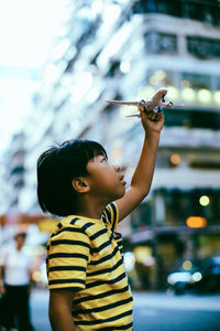 Side view of boy holding model airplane while standing on street in city