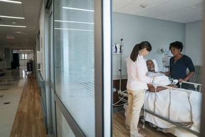 Female doctor talking with senior patient and woman in hospital ward seen through doorway