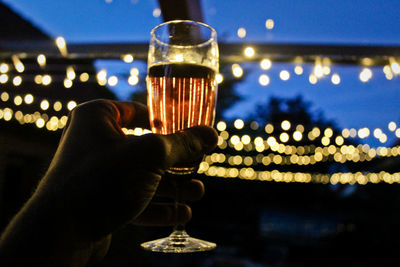 Cropped hand of man holding champagne flute against illuminated lights