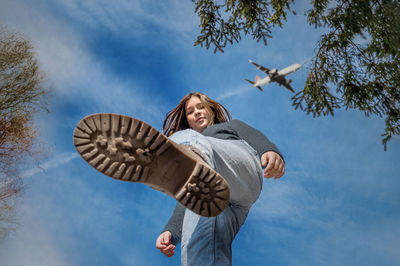 A teenager girl stands with her leg raised outdoors against a blue sky with an airplane. bottom view