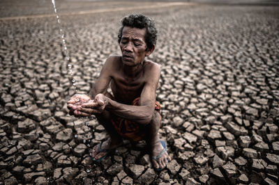 Shirtless man holding falling water while crouching on cracked land