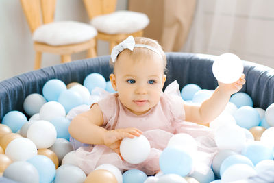 A cute blue-eyed one-year-old baby in a pink dress is bathing in a toy pool with colorful balloons.